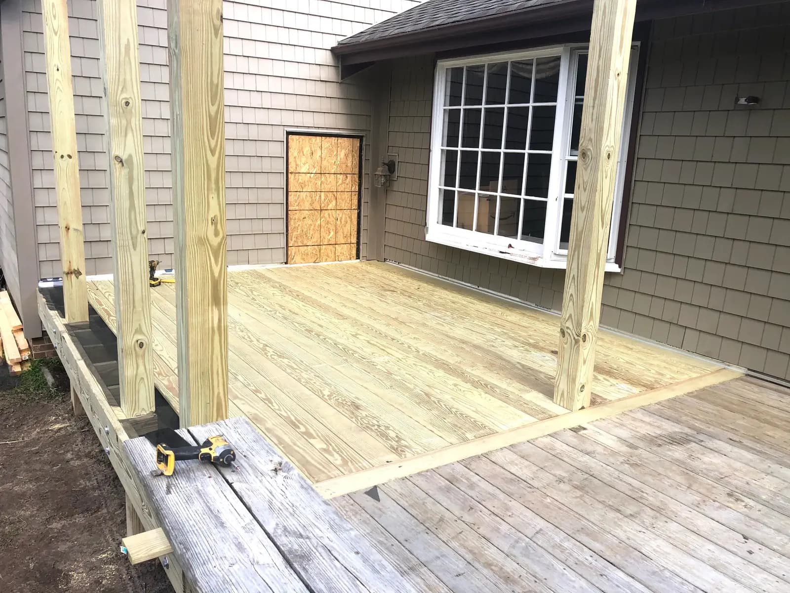 A deck extension in progress: new pressure-treated decking and posts meet the weathered boards of the existing deck against a shingle-sided house.